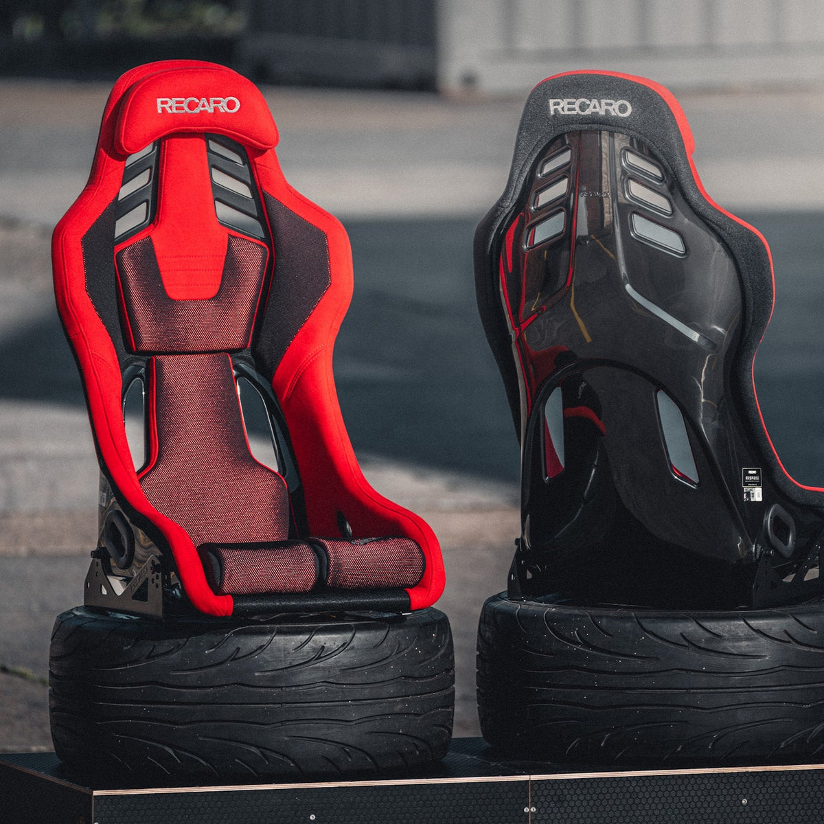 Two racing seats, one red and one black, on wheels with a blurred street background.